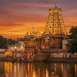 A majestic golden-hued Kanaka Durga Temple illuminated against a vibrant sunset sky, perched atop Indrakeeladri Hill in Vijayawada. The temple's ornate Dravidian architecture is highlighted with intricate details and glowing lights. In the foreground, a serene Krishna River reflects the temple's radiance. Devotees in colorful traditional attire ascend the steps, carrying offerings. Surrounding the temple are lush green trees and floating lotus flowers. The scene is rendered in a style blending hyper-realistic digital art with elements of Madhubani and Pattachitra, featuring rich, deep colors like saffron, maroon, and peacock blue. Soft, ethereal light bathes the entire scene, creating a spiritual and inviting atmosphere.