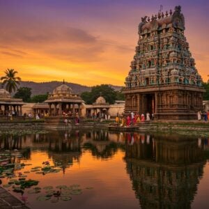 A majestic ancient temple complex in South Indian Dravidian architecture style, illuminated by warm golden light at sunset. The main gopuram of Sri Prasanna Venkateswara Swamy Temple rises prominently, adorned with intricate carvings and colorful sculptures of deities. In the foreground, a serene pond reflects the temple's image, surrounded by blooming lotus flowers. Devotees in traditional attire offer prayers and perform rituals. The sky is a blend of deep orange and purple hues, with hints of stars appearing. Surrounding the temple are lush green hills and coconut trees, emphasizing its picturesque location. The scene is rendered in a style that combines photorealistic details with the ethereal quality of traditional Indian miniature paintings, creating a captivating and spiritually charged atmosphere.