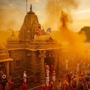 A majestic aerial view of the golden-hued Khandoba Temple in Jejuri during the Bhandara festival, with millions of turmeric particles floating in the air creating a mystical golden atmosphere. The temple architecture features intricate Maratha-style details with stepped pyramidal shikhara, carved pillars, and decorative arches. In the foreground, devotees dressed in traditional Maharashtrian attire (men in white dhoti-kurta and women in nauvari sarees) are throwing turmeric powder skyward. Lord Khandoba appears as a divine manifestation in the golden mist - mounted on a white horse, holding a sword in his right hand and a bowl in his left, wearing a traditional Marathi turban. The scene is illuminated by warm sunset light creating dramatic shadows and highlighting the temple's stone architecture. Sacred flags and bells adorn the temple premises, while traditional drums (dhol-tasha) are visible in the corner. The composition uses a rich color palette of saffron, deep maroon, and gold, with intricate Madhubani-style patterns integrated into the sky. The overall atmosphere is surreal and spiritually uplifting, with lens flares and bokeh effects enhancing the divine ambiance.