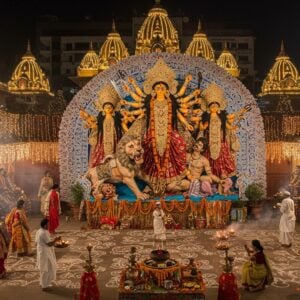 A majestic aerial view of New Delhi Kali Bari temple complex at dusk, illuminated with thousands of warm golden lights and traditional Bengali-style pandal decorations. In the center, a stunning 15-foot-tall traditional Durga idol in classical Bengali style, showing Goddess Durga with 10 arms holding traditional weapons (clockwise: sword, chakra, conch, lotus, arrow, trishul, thunderbolt, bow) mounted on her lion, slaying Mahishasura. The idol features intricate Madhubani-style details, adorned with rich red Benarasi saree, gold jewelry, and a glowing crown. The foreground shows devotees in traditional Bengali attire (women in white-red bordered sarees, men in kurtas) performing dhunuchi dance with incense burners creating mystical smoke patterns. The scene is surrounded by detailed alpana (Bengali floor art) patterns in white, with brass temple bells, marigold garlands, and red hibiscus flowers adding depth. The background features modern Delhi architecture silhouettes merging with traditional temple architecture, creating a harmonious blend of heritage and contemporary elements. The lighting is dramatic with warm golden hues mixed with cool evening blues, creating a spiritually charged atmosphere.