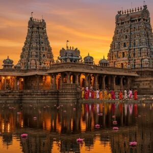 A majestic Hindu temple complex with intricate Dravidian-style gopurams towering against a vibrant sunset sky. The foreground features a serene reflection pool mirroring the ornate architecture. Detailed stone carvings of deities and mythological scenes adorn the walls, with warm golden light illuminating the intricate designs. A group of devotees in colorful traditional Indian attire walk towards the main sanctum, their silhouettes creating a sense of scale. Floating lotus flowers and oil lamps dot the water's surface, adding a mystical ambiance. The scene seamlessly blends ancient architectural elements with a modern, dreamlike quality, using a rich color palette of deep saffron, royal blue, and gold. Soft, glowing halos surround the temple spires, emphasizing their spiritual significance. The overall composition creates a captivating journey through sacred space, inviting viewers to explore further.