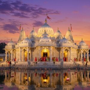 A majestic white marble Swaminarayan temple in Mumbai at twilight, illuminated by warm golden lights, with intricate carved pillars and domes reflecting in a serene lotus pond. The central dome features detailed Indian architectural elements with gold-leafed spires reaching skyward. In the foreground, peaceful devotees in traditional Indian attire (white kurtas and colorful sarees) circumambulate the temple. The scene is enhanced by floating diyas on the water, creating magical reflections. Soft purple and orange clouds frame the temple, while delicate flower petals drift in the gentle breeze. The architecture showcases ornate Gujarati and Rajasthani style carvings with geometric patterns and floral motifs. Ethereal golden light streams through the temple's ornate jharokhas, creating a divine ambiance. The temple is surrounded by perfectly manicured gardens with blooming lotus flowers and fragrant jasmine. Art style combines photorealistic architecture with ethereal lighting and mystical elements, rendered in high detail digital art style with rich textures and atmospheric depth.